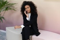 Femme d'affaires noire portant des vêtements formels assis dans le bureau et parlant au téléphone. — Photo de stock