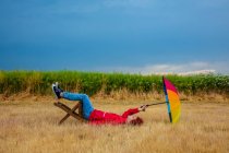 Хлопець з парасолькою сидить на стільці, лежачи на землі на полі — стокове фото