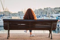 Dos d'une jeune rousse assise sur un banc dans un port de mer — Photo de stock