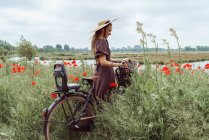 Mulher com bicicleta em pé entre campo de papoilas contra o céu — Fotografia de Stock