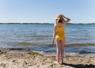 Tween girl in yellow swimsuit and sunhat standing by lake shore. — Stock Photo