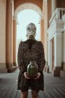 Chica con una máscara de gas militar y un vestido con una planta en sus manos durante la cuarentena, en una ciudad vacía - foto de stock