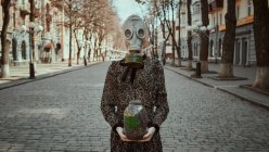 Fille dans un masque à gaz militaire et une robe avec une plante dans ses mains pendant la quarantaine, dans une ville vide — Photo de stock