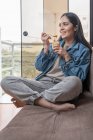 Ritratto verticale di una donna latina sorridente mentre mangia burro di anacardi fatto in casa mentre è seduta su un divano in una casa — Foto stock