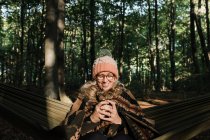 Жінка в капелюсі і окулярах з чашкою кави в лісі . — стокове фото