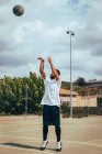 Jovem jogando em uma quadra enquanto atira basquete para cesta — Fotografia de Stock