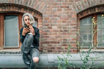 Жінка використовує смартфон, що сидить біля старої цегляної будівлі — стокове фото