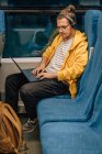 Junger Teenager mit Kopfhörern, fährt mit Laptop im Zug, Programmierer arbeitet aus der Ferne. Vertikale Aufnahme, Porträt eines Reisenden. — Stockfoto