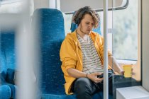 Молодий чоловік, підліток, подорожує в поїзді з навушниками, п'є каву, слухає музику. Знімок стилю з простором для копіювання . — стокове фото