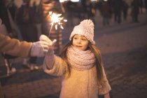 Petite fille décorée de guirlandes vives et tient des étincelles brûlantes sur la place de la ville avec un arbre de Noël — Photo de stock