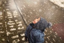 Niño pequeño con ropa azul de invierno mirando al cielo nevando - foto de stock