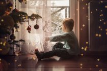 Toddler boy sitting on the floor by the window next to a christm — Stock Photo