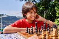 Young pensive boy sitting behind chess board moving one of the pieces. — Stock Photo
