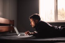 Serious man using laptop computer lying on bed at home — Stock Photo