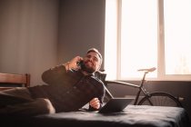 Счастливый человек разговаривает по смартфону лежа на кровати дома — стоковое фото