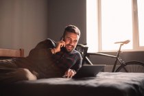 Happy man talking on smart phone while using laptop computer at home — Stock Photo