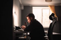 Человек с помощью смартфона и компьютера сидит за столом, работая дома — стоковое фото