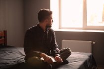 Hombre reflexivo con el ordenador portátil mirando a través de la ventana sentado en la cama en casa - foto de stock