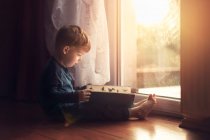 Vista lateral do menino pequeno sentado no chão pela janela e ler — Fotografia de Stock