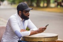 Молодий чоловік сидить за столом у вуличному кафе і дивиться в смартфон. Він у модній білій сорочці та кепці. — стокове фото