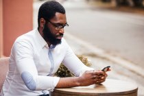 Le jeune homme est assis à une table dans un café de rue et regarde dans le smartphone. Il porte une chemise et une casquette blanches — Photo de stock