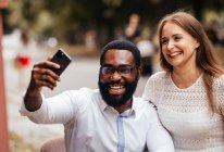 The beautiful young couple of diverse people are taking a photo at a smartphone in the city — Stock Photo