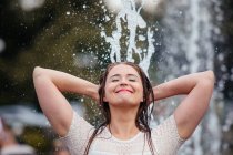 The beautiful young woman in a white knitted dress walks through the fountain in the city. She spreads her arms and laughs — Stock Photo