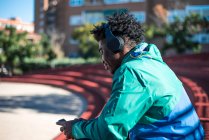 Молодой черный человек расслабляется, слушая музыку и пользуясь своим мобильным телефоном. Сидя в городе. — стоковое фото