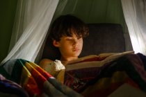 Маленький мальчик лежит на кровати с книгой и смотрит в камеру — стоковое фото