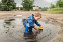 Small blonde boy in blue jacket and blue wellies playing with sh — Stock Photo