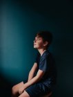 Продуманий молодий хлопчик сидить на табуреті на темно-синій стіні . — стокове фото