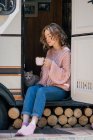 Woman drinking coffee with cat at door of trailer, full length. — Stock Photo