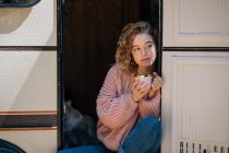 Woman drinking coffee with cat in door of trailer. — Stock Photo
