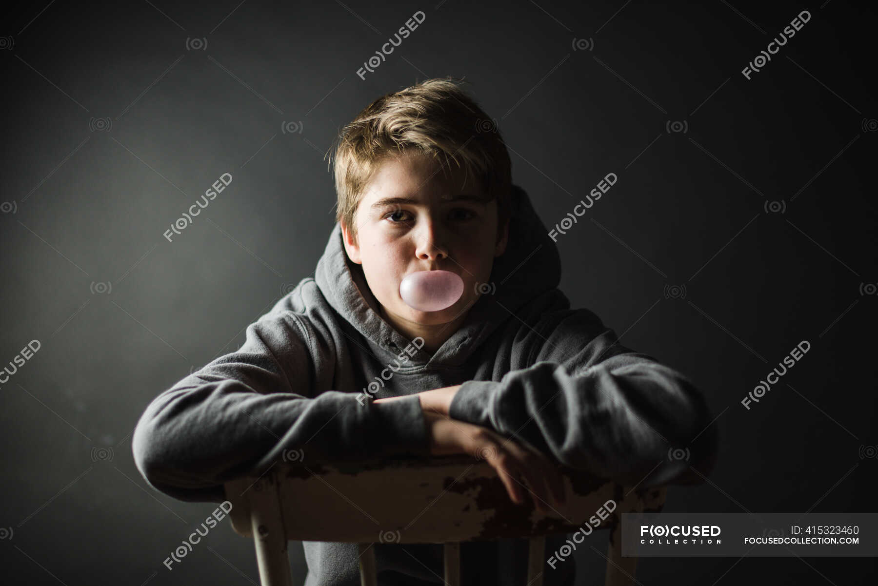 Teen boy in hoodie sitting on a chair in dark room blowing a bubble