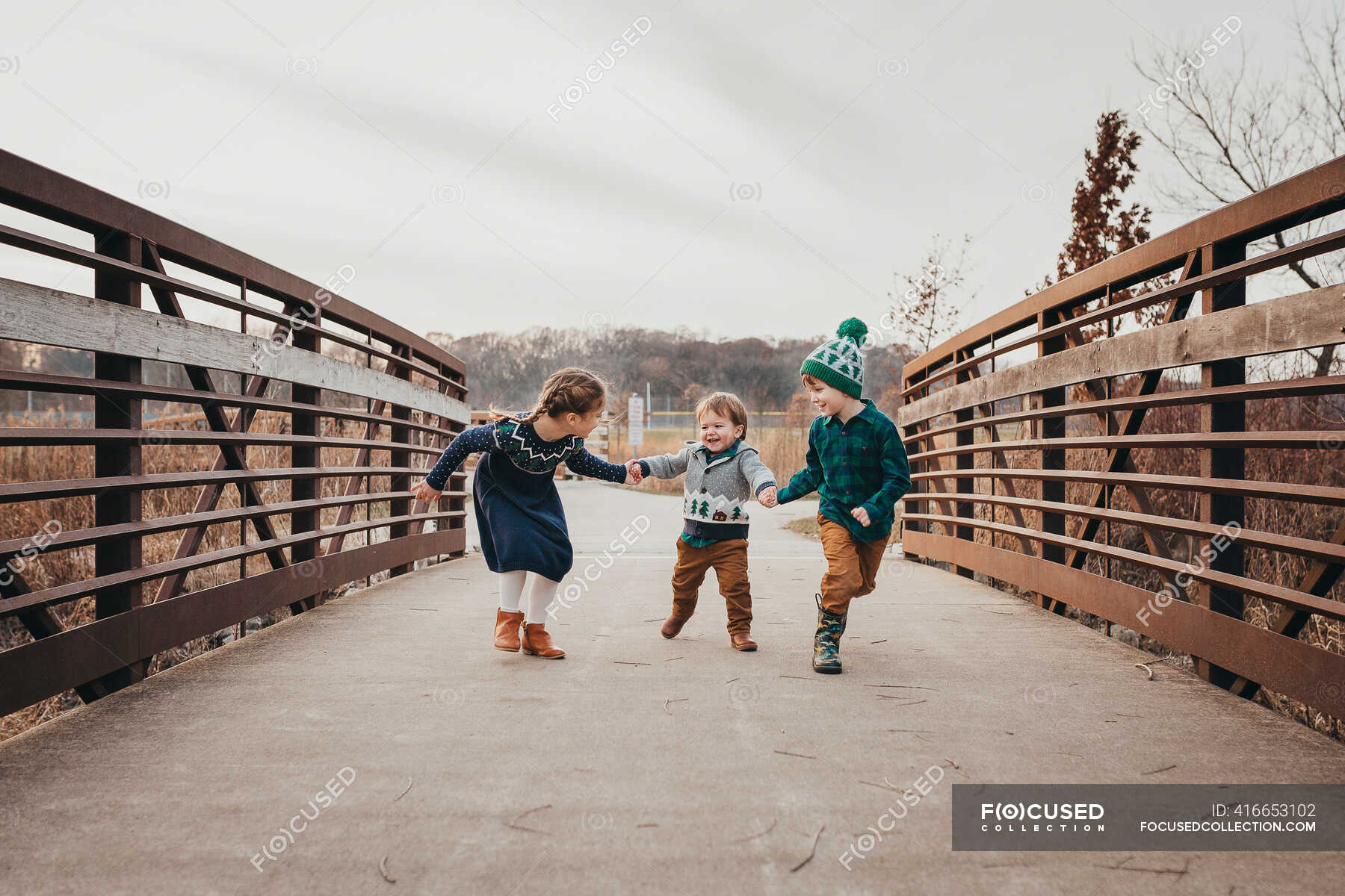 Three siblings laughing holding hands running on bridge — preschool ...