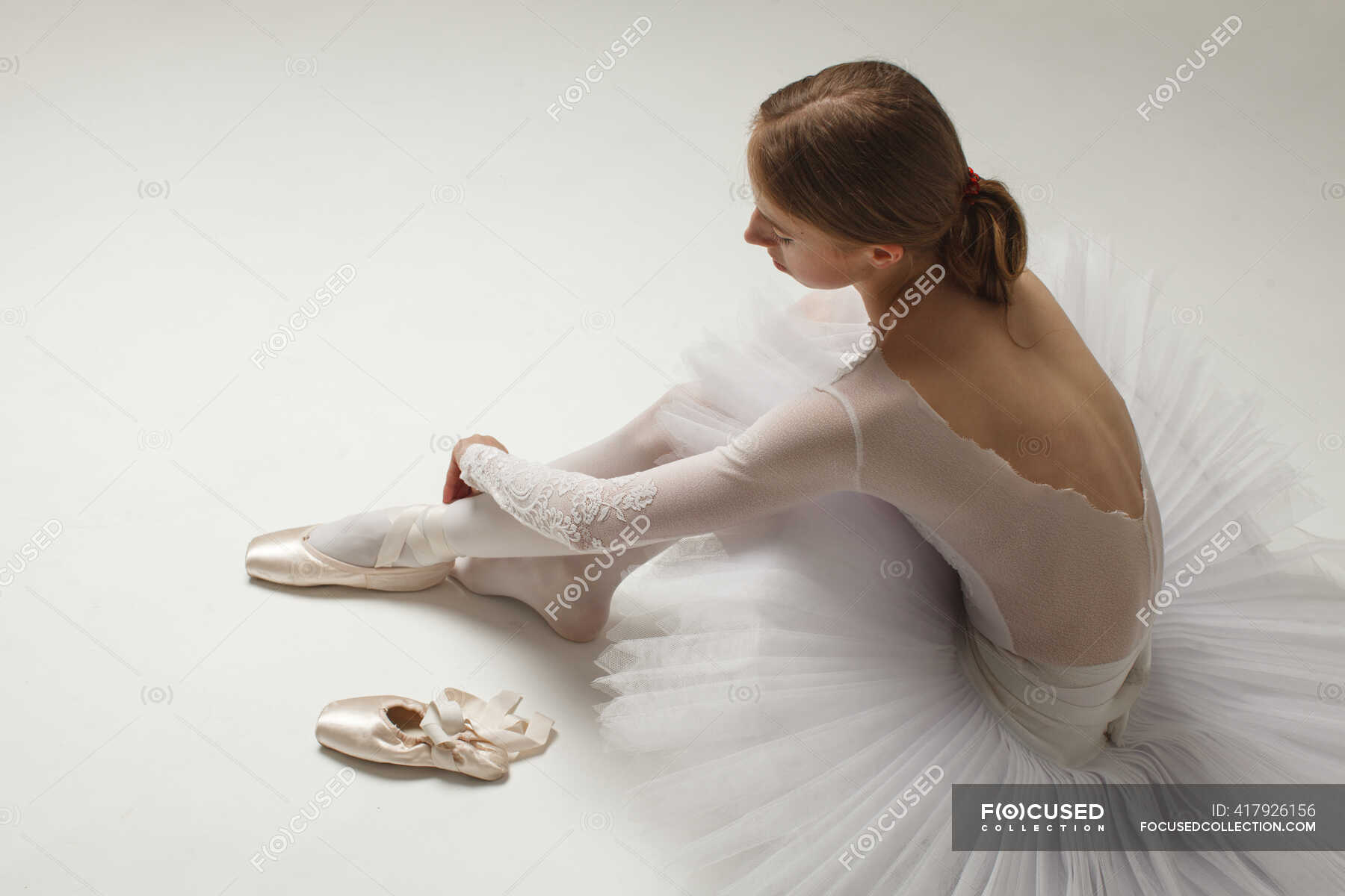 Young ballerina in white ballet dress Putting On Ballet Shoes, sitting on white — beauty, elegance - Stock Photo | #417926156