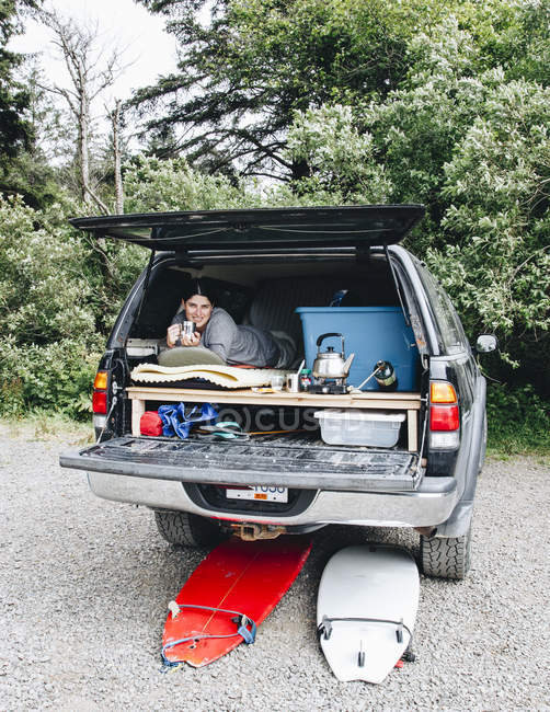 A woman on a surf trip to La Push, Washington lays in a bed in the back of her pickup truck and drinks a coffee to warm up after a surfing session. — Stock Photo
