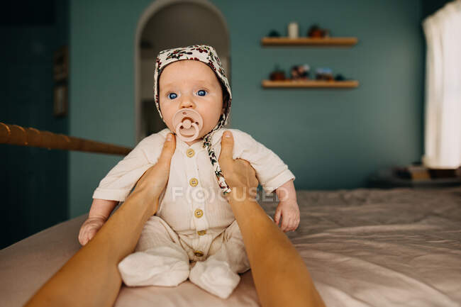 Bebé pequeño en un capó sentado en la cama con la ayuda de las manos de la madre - foto de stock