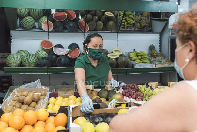 Lojista com uma máscara atende a um cliente no greengrocer — Fotografia de Stock