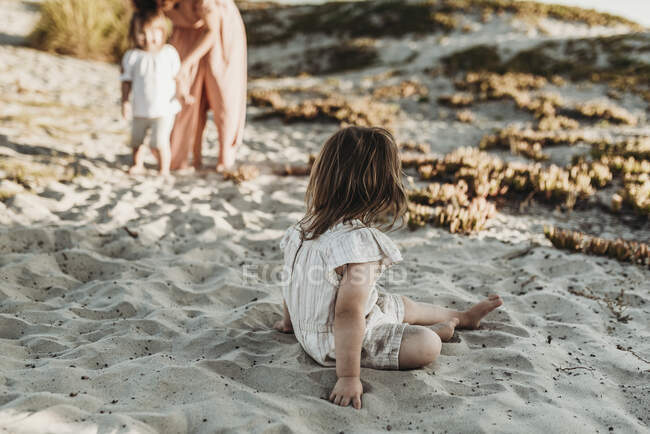 Detrás de la vista de la joven niña sentada en la arena mirando a la familia - foto de stock