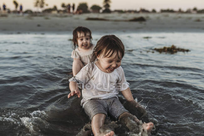 Вид спереди маленьких сестер, сидящих и плещущих в воде на пляже — стоковое фото
