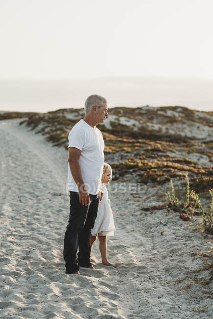 Отец и дошкольница в возрасте стоят в песке на пляже и смотрят в сторону — стоковое фото
