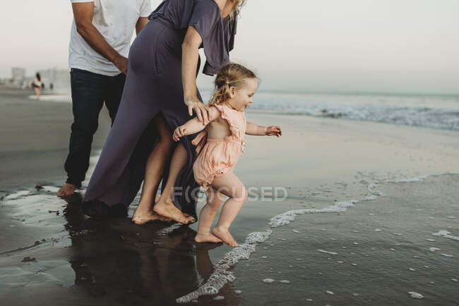 Vista lateral da menina da criança com a família correndo para o oceano ao pôr do sol — Fotografia de Stock