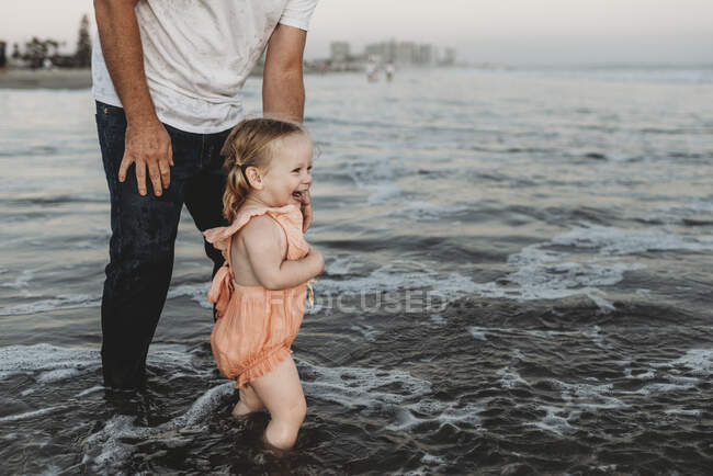 Petite fille éclaboussant dans l'océan avec son père au coucher du soleil — Photo de stock