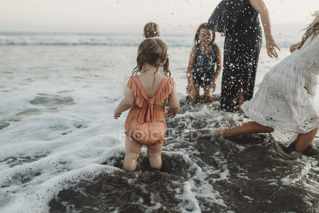 Joven niña chapoteando con hermanas y madre en la playa al atardecer - foto de stock