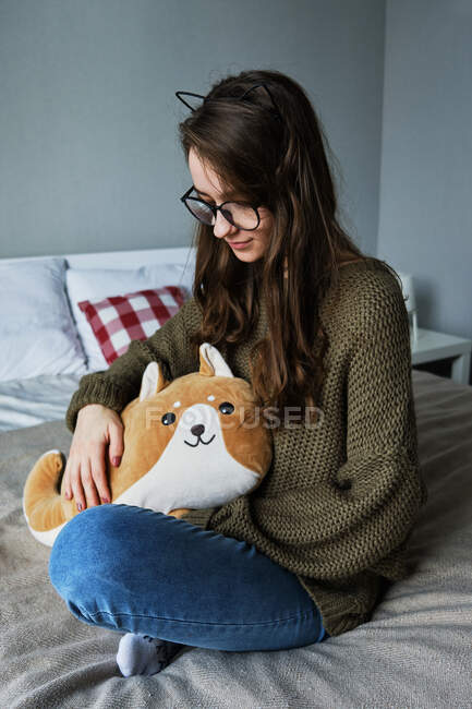Millennial fille aux cheveux bruns dans les oreilles de chat est assis sur un lit avec un jouet — Photo de stock