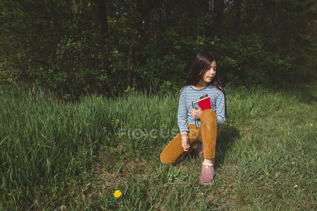 Une fille paisible s'assoit dans l'herbe haute regardant au loin avec un livre dans ses bras — Photo de stock