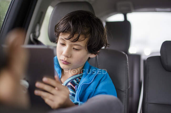 A boy sits in a carseat on a car trip watching a tablet — Stock Photo