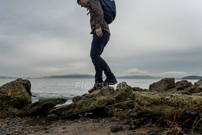 Man with backpack climbs small path of rocks beside bay under gray sky — Stock Photo