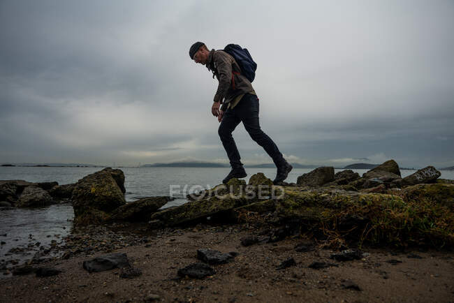 Hiker walks across mossy muddy stones from structure on bayside beach — Stock Photo